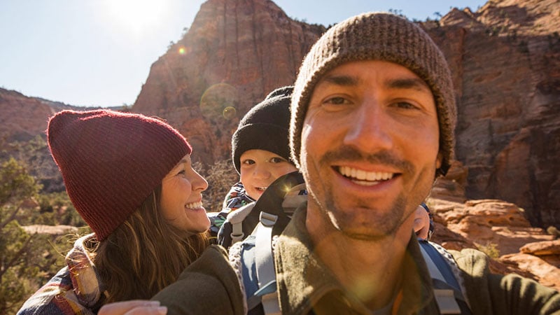 A smiling family of three wearing beanies, hiking through a sunlit canyon with red rock formations, conveying joy and adventure in the outdoors.