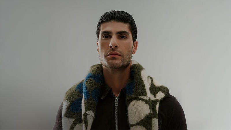 Young man with slicked-back hair wearing a dark zip-up jacket and a patterned scarf against a neutral background, projecting a confident expression.