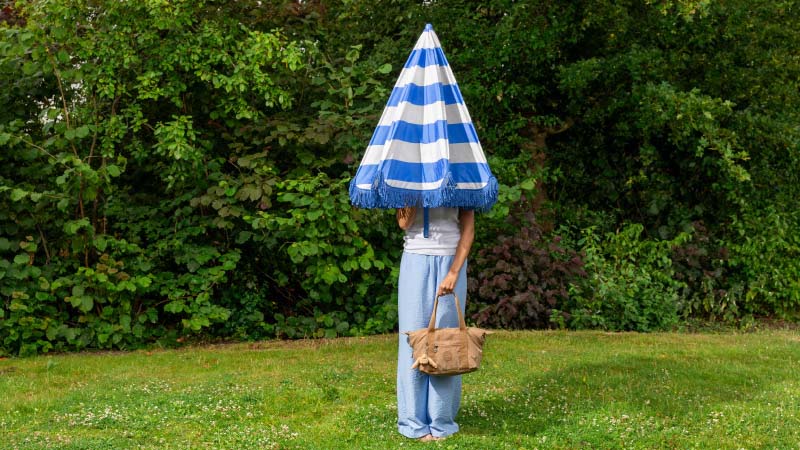 A person stands in a garden, body visible but face obscured by a blue and white umbrella. They hold a brown bag, wearing light blue pants. The scene is quirky and playful.