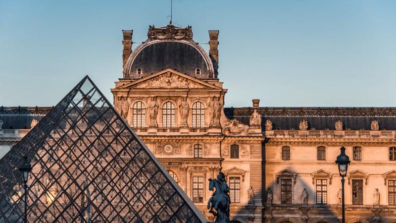 The image shows the Louvre Museum in Paris, featuring the glass pyramid in the foreground and the historic, ornate building facade in the background under a clear sky.