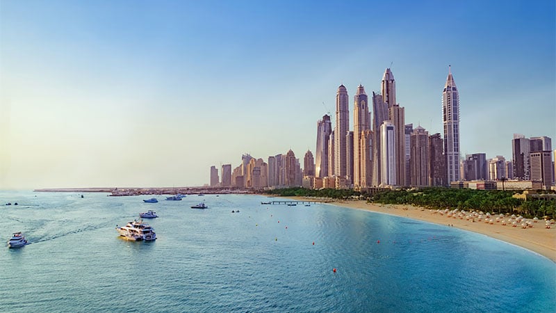 A view of tall modern skyscrapers along a sandy beach with several boats in the blue water under a clear sky.