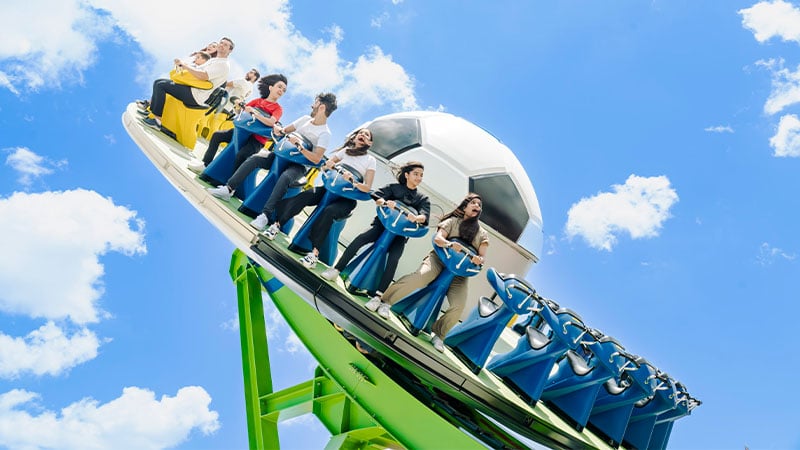 People ride a roller coaster themed with a large soccer ball, set against a bright blue sky with scattered clouds.