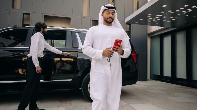 A man in a white kandura walks while checking his red phone. He's next to a black car, with another man opening the vehicle's door. The scene conveys modernity and sophistication.